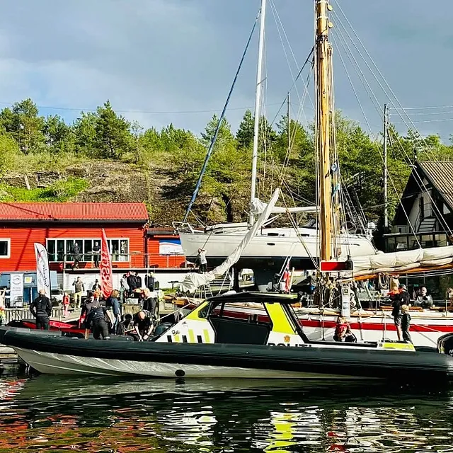 Politibåt og trebåt ligger ved brygga i en livlig havn med folk, flagg og en rød bygning i bakgrunnen.
